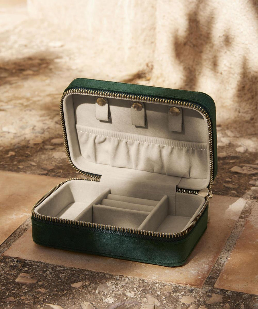 The Bagbase Velvet travel jewellery box, featuring a small green zipped design and light beige interior with multiple compartments and ring holders, is shown open on tiled stone flooring.