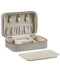 A rectangular beige Bags jewellery box with a gold zip is open to reveal ring rolls, small compartments, elastic pouches, and three press studs inside; its matching lid is set in front.