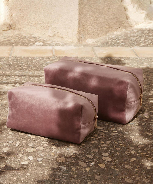 Two Bagbase Velvet toiletry/accessory cases in rectangular mauve with gold zips are placed side by side on a textured stone surface, sunlight casting a shadow in the background.