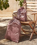 Three Bagbase Velvet gift bags in different sizes are displayed on and beside a wooden folding chair on a stone patio, with leafy branches and a sunlit beige wall in the background.