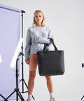 A person with blonde hair stands in a studio setting against a light purple backdrop, holding a large black Bagbase BG721 tote bag. They are dressed in a grey sweatshirt, pink shorts, and sneakers. Studio lights and equipment are visible in the background.