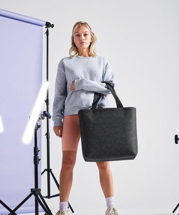 A person with blonde hair stands in a studio setting against a light purple backdrop, holding a large black Bagbase BG721 tote bag. They are dressed in a grey sweatshirt, pink shorts, and sneakers. Studio lights and equipment are visible in the background.