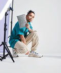 A person with a top knot hairstyle, wearing a teal oversized T-shirt, beige cargo pants, and white sneakers, crouches while holding a gray BG725 notebook by Bagbase. A tripod stand is visible on the left side, set against a plain white background.