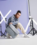 A person wearing a plaid hoodie, light-colored cargo pants, and white sneakers poses in a studio. They are crouching and holding a gray Bagbase BG726 bag. The background features a light purple wall and several lit-up X-shaped and straight light bars on stands.