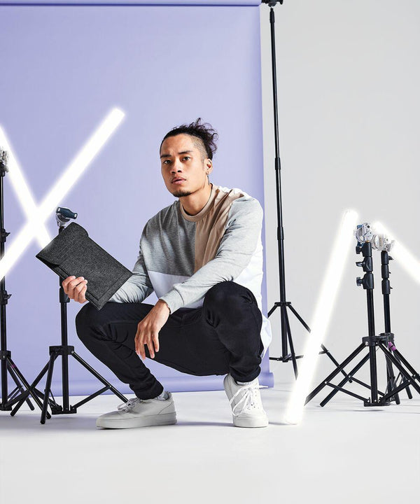 A person with a bun hairstyle kneels on one knee while holding a small dark Bagbase BG727. They wear a color-blocked long-sleeve shirt, black pants, and white sneakers. Behind them is a purple background and multiple studio lights, one of which creates an "X" shape with light tubes.