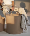 A person dressed in light-colored clothing sits on a chair in the background, while a large beige fabric storage basket from Bagbase, model BG728, is in the foreground. The basket has two handles and contains a light-colored woven textile draped over the side.