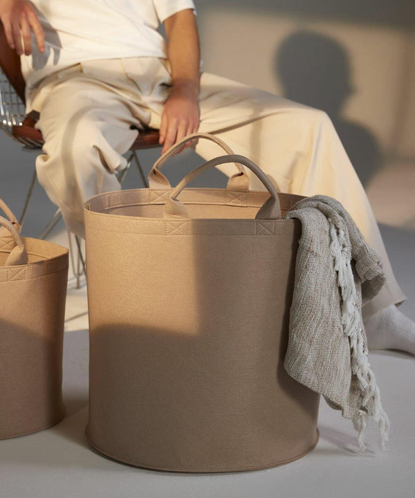 A person dressed in light-colored clothing sits on a chair in the background, while a large beige fabric storage basket from Bagbase, model BG728, is in the foreground. The basket has two handles and contains a light-colored woven textile draped over the side.