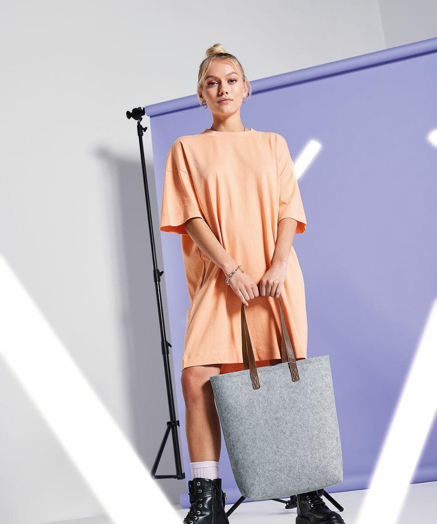 A person stands against a light purple backdrop, wearing an oversized peach-colored T-shirt dress and black combat boots. They hold a large grey Bagbase BG738 with brown handles and have a confident expression. Studio lighting equipment is visible in the background.