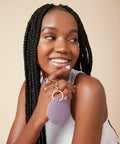 A woman with long braids smiles and looks to the side. She holds a Bagbase BG745 keychain with two rings, close to her face. She is wearing a white sleeveless top and has light purple nail polish. The background is a plain beige color.