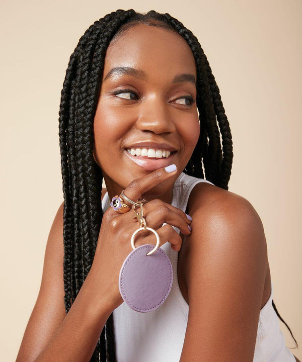 A woman with long braids smiles and looks to the side. She holds a Bagbase BG745 keychain with two rings, close to her face. She is wearing a white sleeveless top and has light purple nail polish. The background is a plain beige color.
