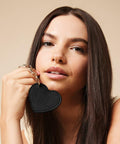 A person with long brown hair is holding a Bagbase BG746 with a gold chain close to their face. The background is a neutral beige color. The person is looking into the camera with a slight smile.