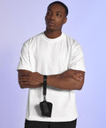 A man stands with arms crossed, dressed in a plain white T-shirt and black trousers. He wears the Bagbase Boutique pouch wristlet on his wrist. The background features a light purple and blue gradient.