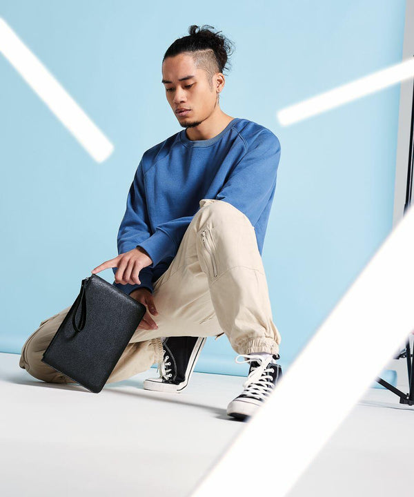 A person with a top knot hairstyle kneels on one knee against a blue background. They wear a blue sweater, beige pants, and black and white high-top sneakers, holding a Bagbase BG750. White light streaks cross the foreground.