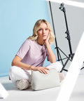 A person with blonde hair, wearing a light purple T-shirt and white pants, is sitting on the floor against a blue and white background. They are holding a Bagbase BG751 handbag. A tripod with lighting equipment is visible in the background, and bright white light streaks intersect the image.