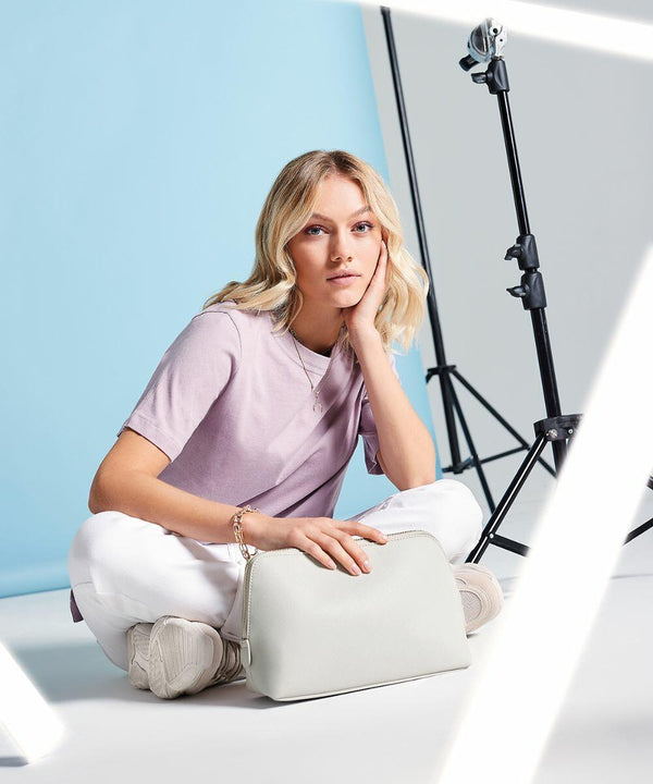 A person with blonde hair, wearing a light purple T-shirt and white pants, is sitting on the floor against a blue and white background. They are holding a Bagbase BG751 handbag. A tripod with lighting equipment is visible in the background, and bright white light streaks intersect the image.
