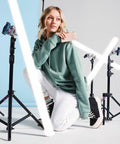 A woman with blonde hair, wearing a green hoodie and white pants, kneels on one knee with light tubes and tripods around her. She looks off to the side and holds a **Bagbase BG754** in her left hand. The background is a mix of blue and white.