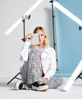 A woman sits cross-legged in a photoshoot setup, holding a closed BG755 from Bagbase over one eye. She wears a white jacket, floral pants, and black platform shoes. Background elements include blue paper, studio lighting, and cameras on tripods.
