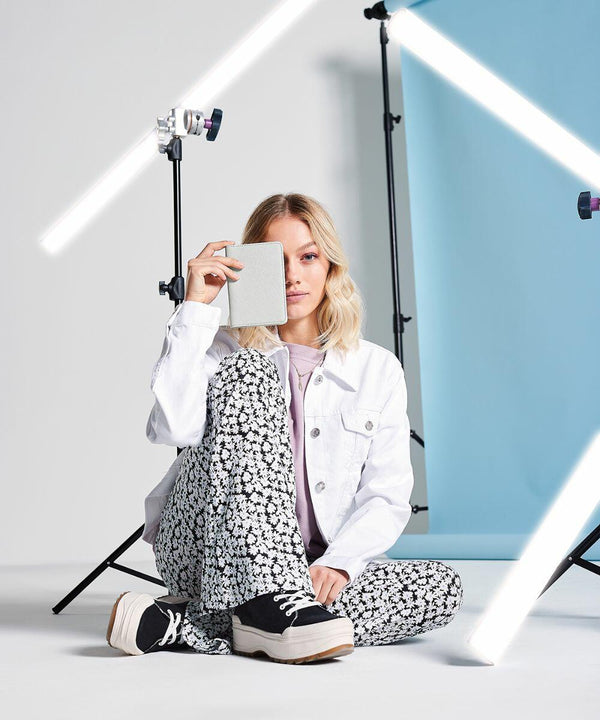 A woman sits cross-legged in a photoshoot setup, holding a closed BG755 from Bagbase over one eye. She wears a white jacket, floral pants, and black platform shoes. Background elements include blue paper, studio lighting, and cameras on tripods.