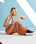 A person with blonde hair sits on the floor against a light blue backdrop, wearing a white tank top, orange wide-leg pants, and black and white sneakers. They hold a gray BG756 clutch bag by Bagbase and pose with a relaxed expression. Sunlight and shadows create diagonal lines.
