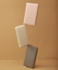 Three rectangular zippered wallets by Bagbase, model BG757, in beige, light pink, and gray are arranged diagonally against a plain brown background. The wallets are standing on their edges, seemingly balancing on one another.