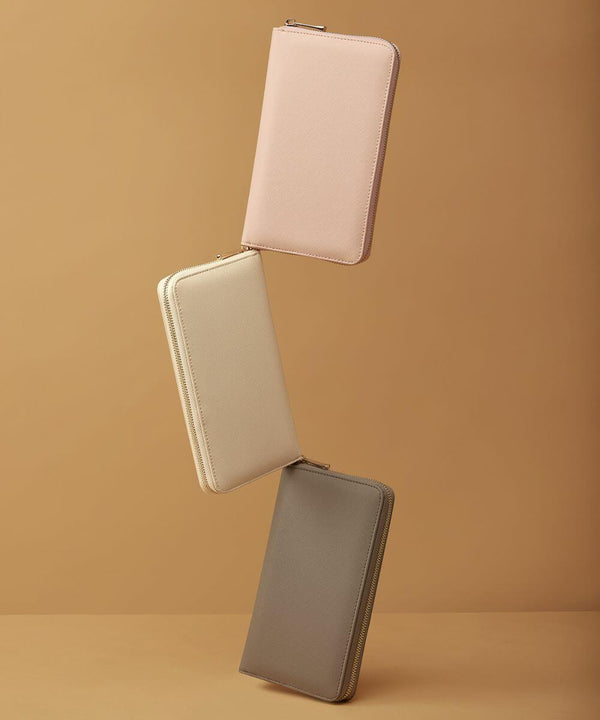 Three rectangular zippered wallets by Bagbase, model BG757, in beige, light pink, and gray are arranged diagonally against a plain brown background. The wallets are standing on their edges, seemingly balancing on one another.