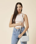 A woman stands against a beige background, dressed in a pale pink sleeveless top and light blue jeans. She holds a Bagbase BG758 crossbody bag in her right hand while her left hand rests on the strap. She has long, straight hair and wears a bracelet on her right wrist.
