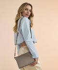 A woman with long, wavy blonde hair is posing against a peach-colored background. She is wearing a light blue, cropped button-up shirt, beige pants, and carrying a gray Bagbase BG759 shoulder bag. She faces slightly to the left with a confident expression.