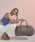 A woman with shoulder-length blonde hair is squatting on the ground. She is wearing a blue sleeveless top, white pants, and yellow heeled sandals. She is holding a large gray Bagbase BG760 handbag with both hands, against a soft pink background.
