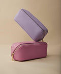 Two rectangular zippered pouches, the BG761 from Bagbase, are shown in a minimalist setting with a beige background. The top pouch is light purple and placed at an angle on top of the bottom pink pouch. Both pouches have gold zippers and a textured appearance.