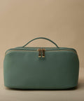 A teal, rectangular travel toiletry bag with a textured surface is displayed against a beige background. The Bagbase BG762 features a handle on top and a gold zipper with two pulls in the center.