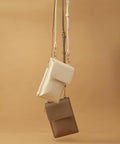 Image featuring two Bagbase BG767 crossbody bags hanging against a warm beige background. The upper Bagbase BG767 is a light beige color, while the lower Bagbase BG767 is a darker taupe shade. Both Bagbase BG767 bags have a slim, rectangular shape with zippered closures and adjustable straps.