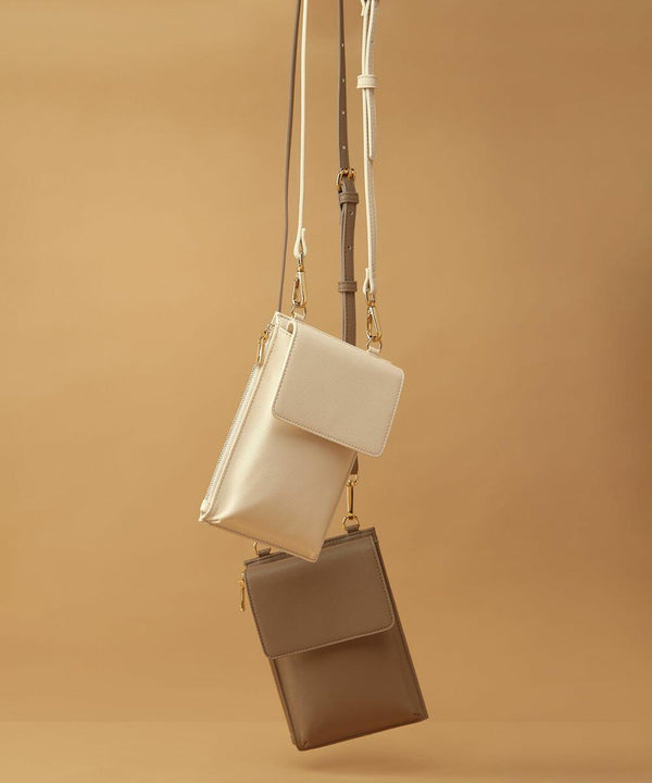 Image featuring two Bagbase BG767 crossbody bags hanging against a warm beige background. The upper Bagbase BG767 is a light beige color, while the lower Bagbase BG767 is a darker taupe shade. Both Bagbase BG767 bags have a slim, rectangular shape with zippered closures and adjustable straps.
