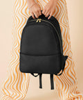A person holding a black Bagbase BG768 down in front of them with both hands. The person is wearing pants with orange and white wavy patterns. The background is a plain beige color. The Bagbase BG768 has a gold zipper and appears to be made of a smooth material.