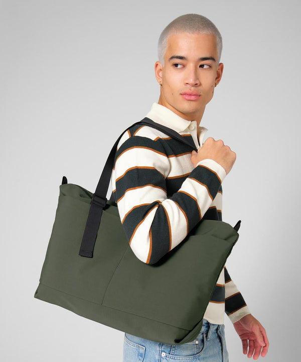 A person with short, fair hair wears a striped long-sleeved shirt and blue jeans, carrying the Bagbase Minimal lifestyle tote over their shoulder, looking to the side against a plain grey background.