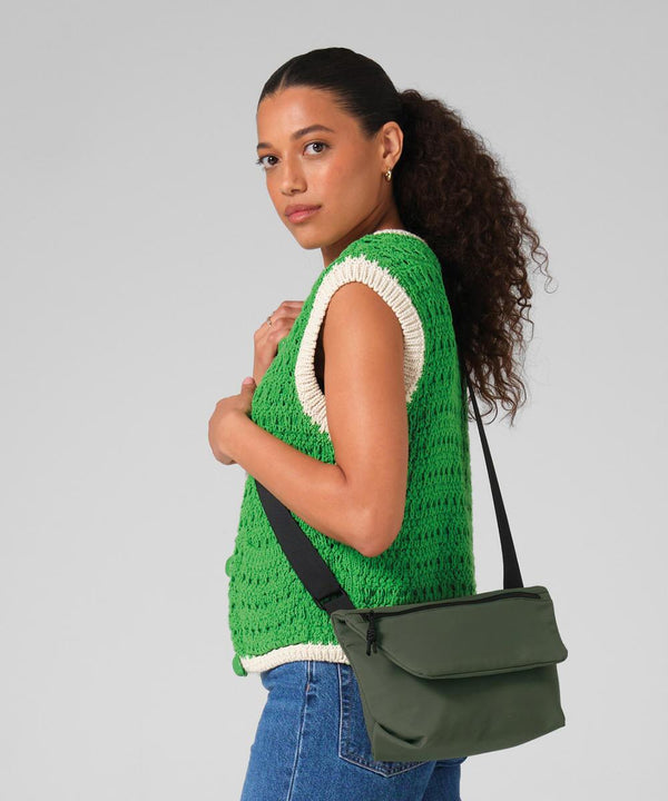 A woman with curly hair in a ponytail wears a green knitted waistcoat, white shirt, and blue jeans. She holds the strap of the Bagbase Minimal lifestyle mini messenger and looks at the camera against a plain grey background.