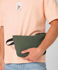 A person in a light pink T-shirt with a postage stamp print and blue jeans is holding the Bagbase Minimal lifestyle pouch, a green zippered accessory with a black wrist strap.