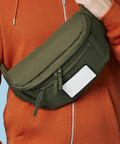 A person in an orange hoodie is wearing an olive green Bagbase BG841 crossbody bag with a front zippered pocket and a blank white rectangular label. The bag has multiple zippers and appears to be made of durable fabric. Only the torso and partial arm of the wearer are visible.