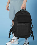 A person dressed in light grey shorts, white socks, and white athletic shoes with orange details is holding a large, black Bagbase BG850 tactical backpack. The background is a solid light blue. The person is holding the Bagbase BG850 by its top handle.