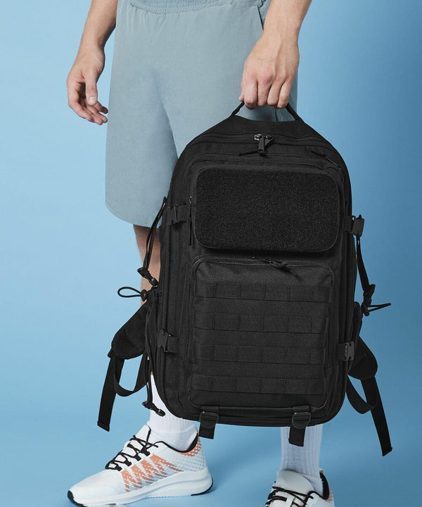 A person dressed in light grey shorts, white socks, and white athletic shoes with orange details is holding a large, black Bagbase BG850 tactical backpack. The background is a solid light blue. The person is holding the Bagbase BG850 by its top handle.