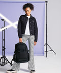 A person with curly hair stands next to a purple backdrop and studio lights, wearing a black denim jacket, white shirt, patterned flared pants, and black sneakers. They hold a large Bagbase BG853 in one hand and look directly at the camera.