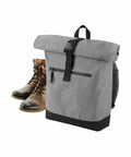 A pair of worn brown leather lace-up boots is placed near a grey Bagbase BG855 with black zipper and buckle closures against a white background. The Bagbase BG855 has a front zip pocket and a black bottom.