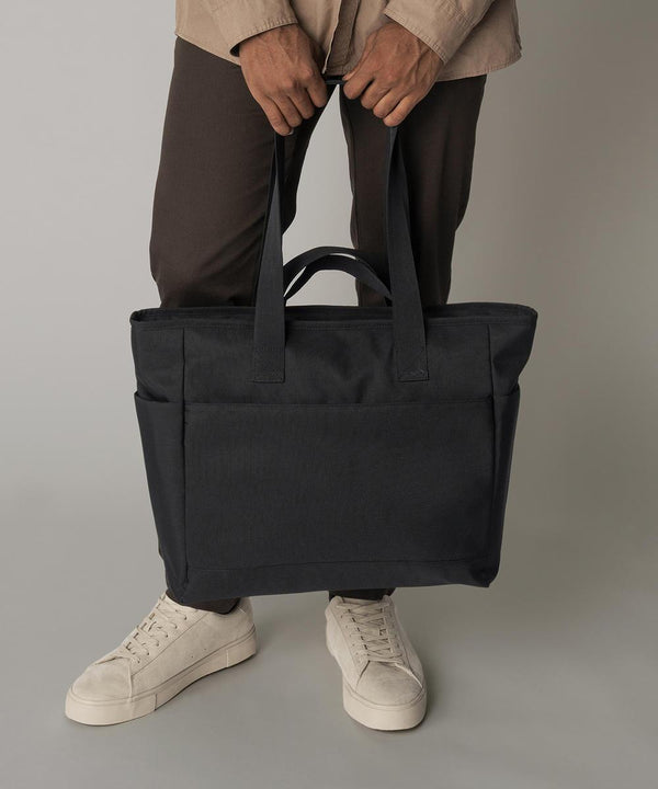 A person stands on a neutral background wearing brown trousers, a beige shirt, and white trainers, holding the Bagbase Simplicity tote, a large black bag, by its handles.