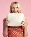 A woman with straight blonde hair holds the large cream-coloured Sundae accessory case by Bagbase in front of her mouth. Dressed in a sleeveless red checked dress with light blue nails, she stands against a pink background.