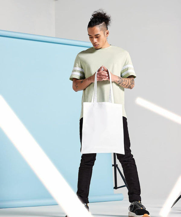 Person with a bun hairstyle wearing a light green shirt and black pants holds a white Bagbase BG901 tote bag. Light-blue backdrop and standing lights are visible in the background.