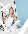 A person with long, light-colored hair styled in a high ponytail is sitting cross-legged on the floor. They hold a Bagbase BG915 and are surrounded by studio lighting equipment against a backdrop of white and light blue panels.
