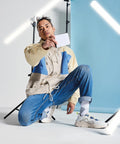 A person with long blonde hair, dressed in a light pink shirt and light blue jeans, crouches while holding a white Bagbase BG960 with a black zipper. The background is a smooth blue, and there are white fluorescent light tubes creating a geometric pattern around them.