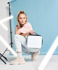 A woman squats, holding a Bagbase BG960 against a light blue backdrop with fluorescent lights around her.