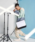 A person in casual attire poses in a studio with a blue background, holding an empty Bagbase BG965 bag while surrounded by studio lighting equipment.