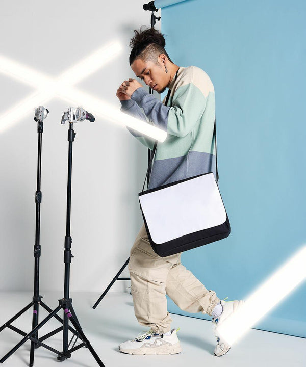 A person in casual attire poses in a studio with a blue background, holding an empty Bagbase BG965 bag while surrounded by studio lighting equipment.