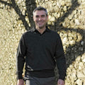 A smiling man with short gray hair is walking outdoors on a sunny day. He is wearing a dark gray Balmoral Lambswool sweater and pants, and the background shows a stone wall with shadows of tree branches cast on it.